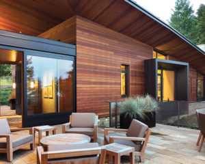 A modern cedarwood house with a patio and forest surrounding it. On the patio, there are two tables for people to sit at. 