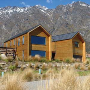 This image depicts two structures with pine siding. There are mountains in the background of the image with snow on the caps. 