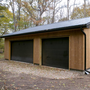 This image depicts a garage with pine siding. There are two garage doors closed and leaves are scattered on the ground.