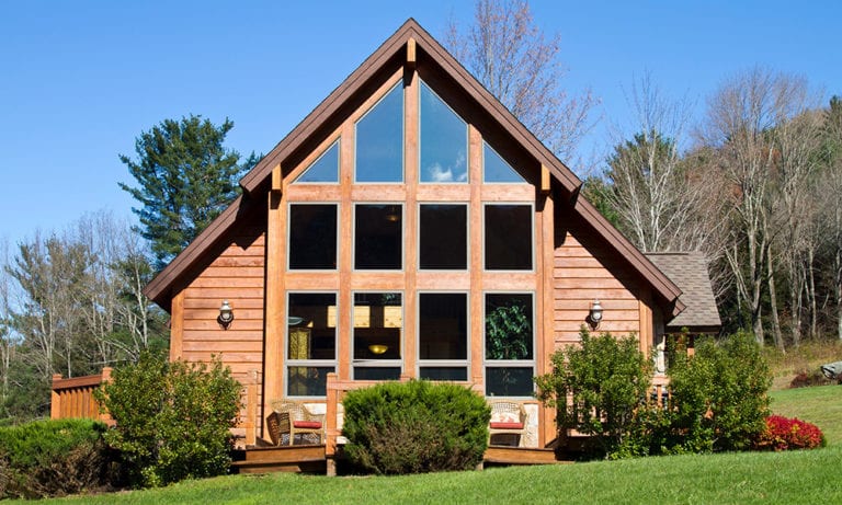 An A-line roofed house with glass paneling. In the photo's foreground is a well-manicured lawn and bushes.