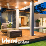 This covered wood deck is installed next to a pool. A chair, coffee table and ottoman are sitting on the deck.