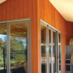 The picture depicts an interior corner of a home with orange wood and two glass sliding doors on either side of the corner. There is a grill against a mosaic wall in one corner and a picnic style table in the other.