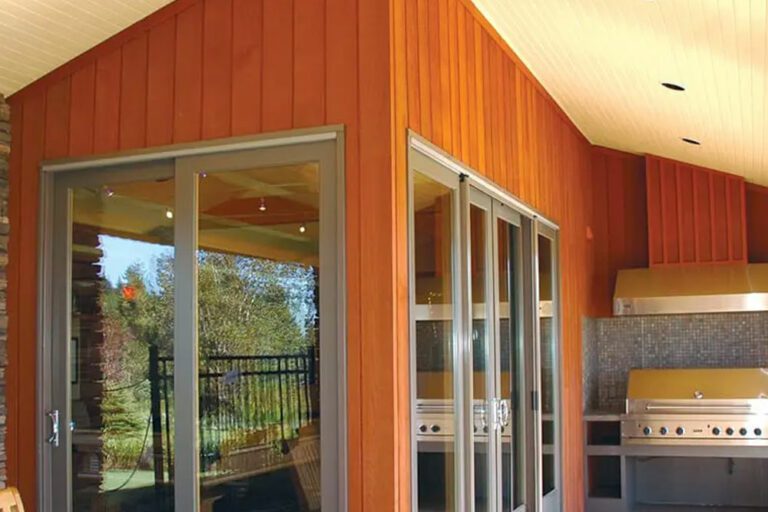 The picture depicts an interior corner of a home with orange wood and two glass sliding doors on either side of the corner. There is a grill against a mosaic wall in one corner and a picnic style table in the other.
