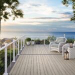 A beach-side deck with light-colored wooden floors, white railings, some potted plants and 2 lounge chairs