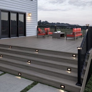 This image depicts a house with an elevated deck design. There is a seating area with red chair and sofa cushions. There is a black railing system on the deck, along with an integrated lighting system on the steps.