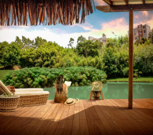 This image depicts two people sitting at the edge of a wooden deck overlooking a pond and lush greenery. One person is partially in the water and the other person is reading a book with their feet in the water. There are two sunbeds and a round coffee table next to them.