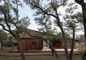 This image depicts a rustic, two-story building with a corrugated metal roof and red cedar siding surrounded by oak trees with a concrete pathway leading up to it. 