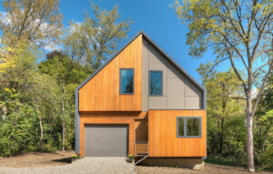 This image depicts a modern house with a steep-pitched roof and light wood paneling surrounded by trees.