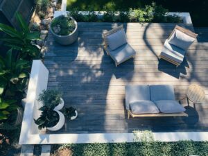 This image depicts an aerial view of a wooden deck. The deck has a set of two armchairs, a loveseat, a small round side table and various potted plants on it.