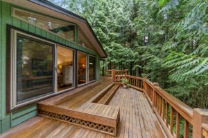 This image depicts an elevated wooden deck off of a green cabin with large windows. The home is surrounded by trees.