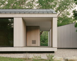 This image depicts a modern wooden building with large glass windows, surrounded by trees and gravel landscaping.