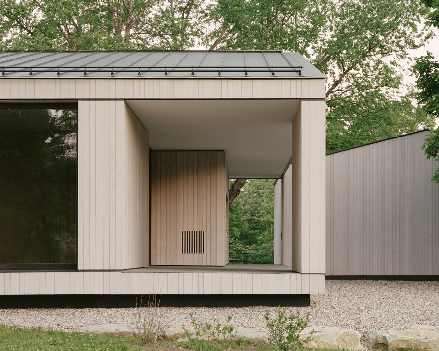 This image depicts a modern wooden building with large glass windows, surrounded by trees and gravel landscaping.