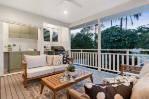 This image depicts a covered deck with an outdoor kitchen and grill. There is a sitting area with two couches and a coffee table between them. A white fan is on the ceiling and white railings overlook some trees.