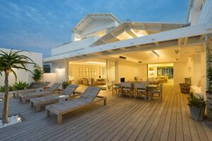 This image depicts a ground-level deck design on a white house. There is a large outdoor lounge and eating area.